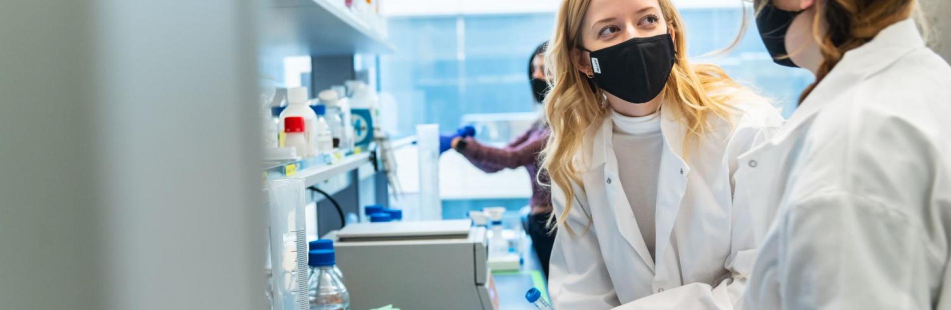 Two Science students in lab coats and masks chatting in the science lab