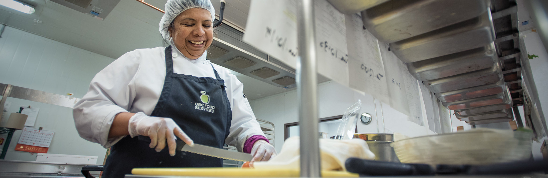 Food Services chef wearing a foodsafe cap slices a tomato with a serrated bread knife on a cutting board