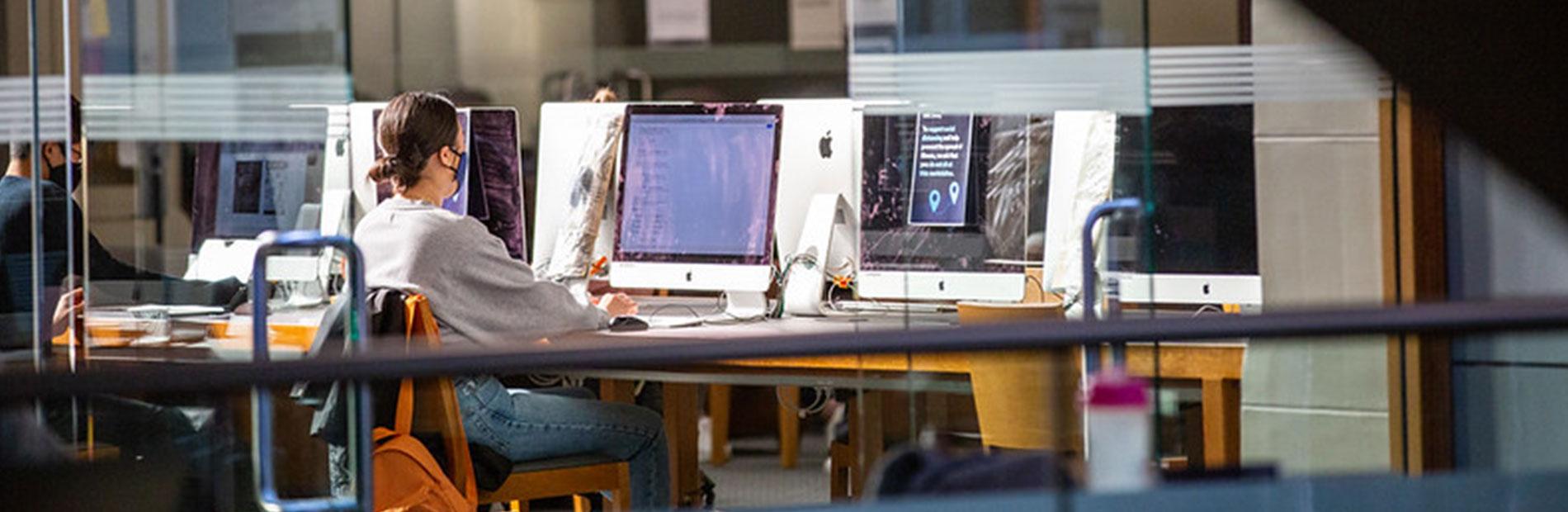 Student wearing a face mask watches a computer screen in the computer lab.