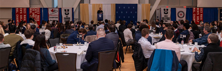 Student-athletes, coaches, alumni, donors, deans, and others sit at round tables at the breakfast event, listening to speaker who stands at a podium.